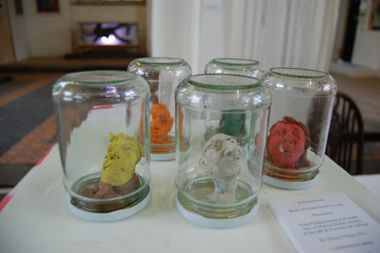Five upside-down jam jars, each containing a plasticine busts. The models are all friends or family of Norwich Dandy Eloise O'Hare. The plasticine heads each use a single colour different from each other. The colours are yellow, white, red, orange and green. In the distant background you can just about see the Damian Hurst tank.