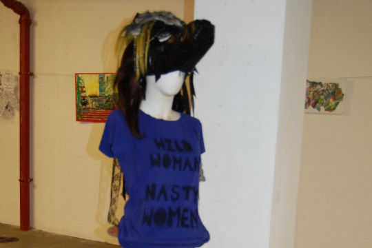 A wolf's head mask made by artist Hayley Hare modelled on a mannequin with a deep blue tee-shirt. The figure was set up on three wooden cable storage reels stacked on top of each other next to one of the large concrete support posts that run throughout the gallery space.