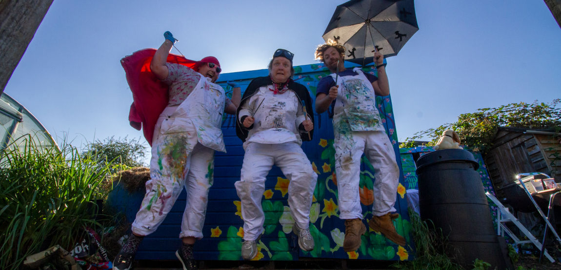 A photo of Chrissy Sabberton, Eloise O'Hare and Dugald Ferguson jumping in front of a colourful shed painted with flowers. The shed was painted by the Norwich Dandies as part of The Great Dandy Mural Project in 2021.