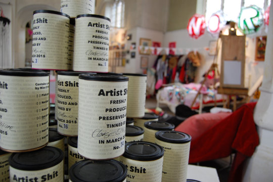 A stack of cans containing Artist Shit. Each can reads that it is 'freshly produced, preserved and tinned by the artist, and each can has its own batch number. In the background is the Dandifest Dress, Paint and Pose Like a Dandy workshop station.