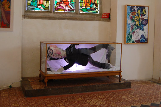 This photo sets the scene for a section of the wider exhibition at St Margaret's Church of Art in Norwich. In the background is a large stained glass window showing religious scenes with the light shining though. Directly underneath it is Eloise O'Hare's Damien Hirst installation. On the wall to the right is a Sportsflower painting by Dugald Ferguson. To the left is an abstracted figurative oil painting in earthern colours depicting a woman in robes. The floor is tiled with old, decorative church flooring in ochre, red brown and black around the floor tile border.
