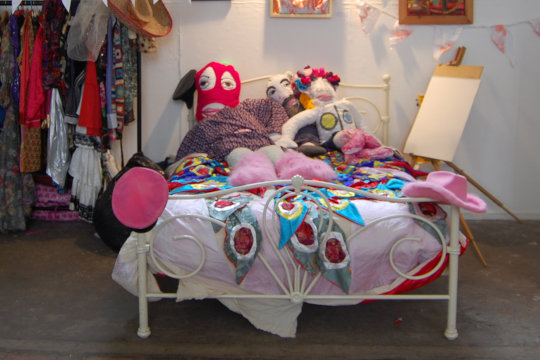 Dugald Ferguson's performance installation 'Our Political Home Life'. The scene features a bed with three of Eloise O'Hare's life-sized textile dolls: a Pussy Riot member and John Lennon and Yoko Ono. To the left is the Dandy costume collection that is used for our 'Dress, Paint and Pose Like a Dandy' workshops. Just above the costumes are a couple of Christina Sabberton's pink portraits of feminist icons. Behind the bed on the wall are a series of expressive mixed media textile paintings and above these are a few of Vince Laws' 'Umbrellas of Love', each with a stencil sprayed letter to spell different words all around the exhibition space.