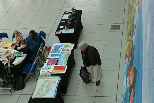 A section of the Dandies’ pop-up Fantabulosa exhibition, showing the workshop area and art shop at the Forum. On the right is one of three large vinyl printed of paintings hanging from the railing of the upper balcony running around the circumference of The Forum. The workshop has a full table of participates drawing and colouring and the foreground shop area are watercolour paintings by Eloise O’Hare.