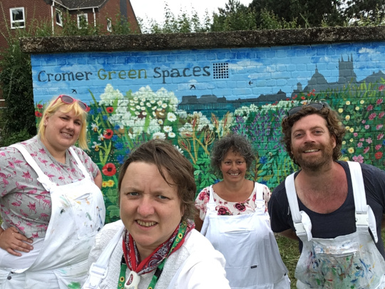 Chrissy, Eloise, Beki and Dugald taking a selfie in front of the finished mural, with paint splatters all over their white overalls.