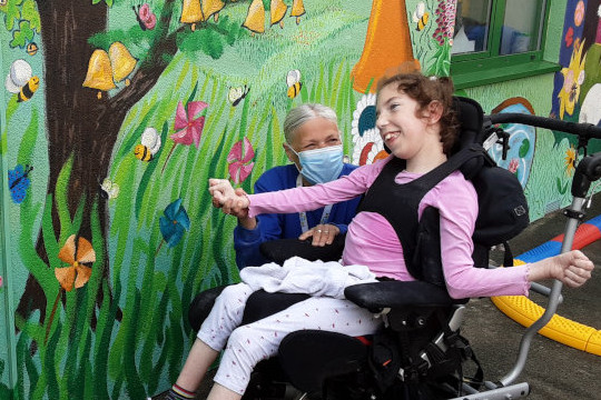 Here we see one of the day users of the Hamlet Charity in her black electric wheelchair looking at a section of the mural with a big smile on her face and her arms held out wide like she wants to hug it. The section of the mural she is responding to has beach windmill flowers in the long grass, an oak tree that is covered in golden bells ringing, an old piano that is covered in flowers on a vine and just behind her an upside down horn that is sprouting blossom. All around there are busy bees gathering nectar.