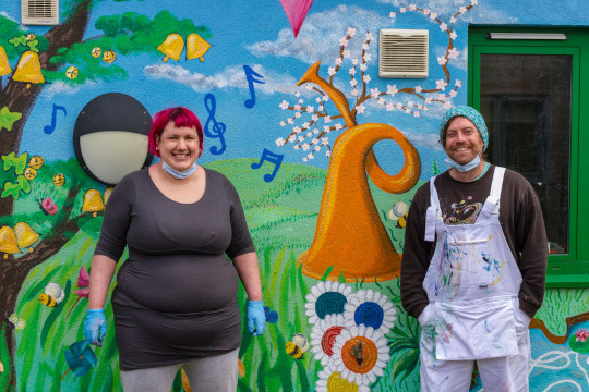 In front of the middle section of the mural we have a mid-length body shot of Chrissy and Dugald having just finished painting the mural. Chrissy’s hair is short and a bright fire engine red. She is wearing a black dress top with grey leggings and blue protective gloves. Dugald has on his warm wooly hat he knitted in turquoise, teal and white yarn. He has white but very paint splattered dungaree overalls covering a black sweatshirt. On the mural between them we see the upside down horn sprouting blossoms and fisher price toys inspired, different coloured records that are part record part daisy growing out the ground.