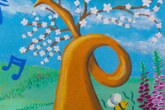 Here we have a detail shot of a section of the mural. The main focus is the big upside down orangey gold horn that is sprouting white blossom. In the sky around and behind it we see dark blue musical notes dancing through the air. To the right of the horn are painted foxglove flowers in a different pink tones. In front of the horn we can see a couple of the colourful record flowers. The horn is sat upside down on the meadow grass and there is a bee buzzing around it looking for nectar.