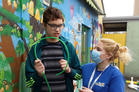 Here we see one of the Hamlet Charity users’ and their carer right next to the new mural. The child is holding a long, thin, green plastic thing that can bend with both of his hands. He is wearing a dark turquoise hoodie top and black and grey striped t-shirt with grey trousers. His carer is wearing the Hamlet uniform which is a cobalt blue t-shirt with black trousers. The blue of the t-shirt tones very nicely with the lighter blue of the mural’s sky in the background. They are both standing nearest the oak trees covered in golden bells. Near their knees are some beach windmill flowers which were inspired by some of the kids drawing them in their art classes.