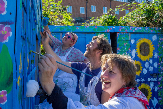 A photo of Eloise, Dugald and Chrissy working on a large outdoors Mural. They are painting flowers on a large blue shed.