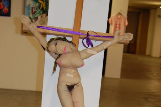 A mixed media installation by Eloise O'Hare of a textile female puppet doll, naked and nailed to a 1.5 metre tall crucifix. In front of the doll is a low table and seat with a typewriter on top. The audience can load strips of paper provided into the typewriter and write abusive, sexist comments on the paper strip and then pin the paper anywhere they like on the puppet doll.