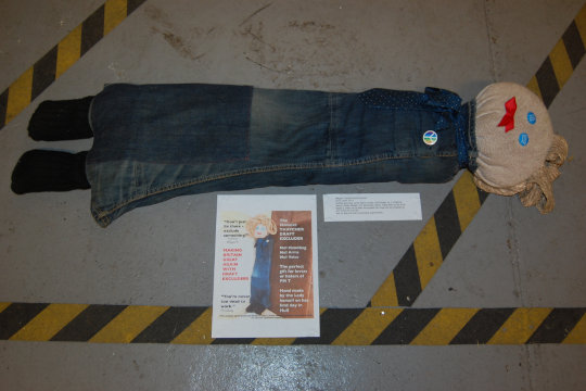 A Margaret Thatcher Draft Excluder by Vince Laws. It is made using denim, socks and wool with trimmings, including blank blue eyes and bright red lips made using an AIDS remembrance ribbon. It is exhibited on the floor with accompanying propaganda style information reading: 'Don’t just lie there - exclude something!', 'Making Britain great again with draft excluders' and 'You're never too dead to work'.